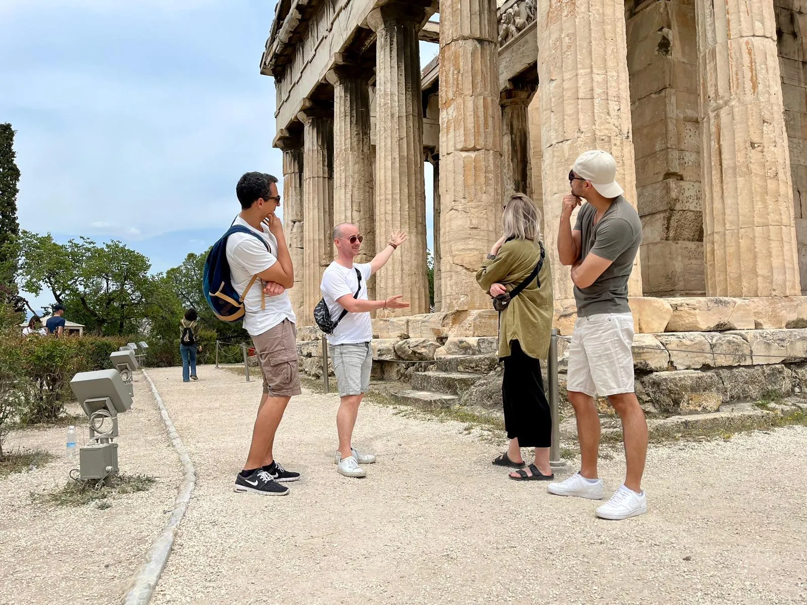 Stefanos als Reiseführer in Griechenland — erklärt Freunden einen Ort auf einem sonnenbeschienenen Steinplatte.